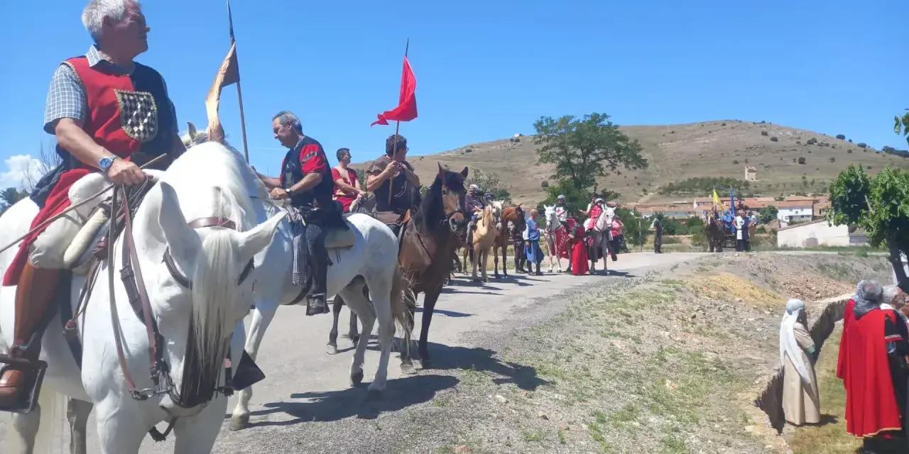 Un desfile ecuestre con trajes históricos, quizás recreando la gesta de el poyo del cid, avanza por un camino rural bajo un cielo azul.