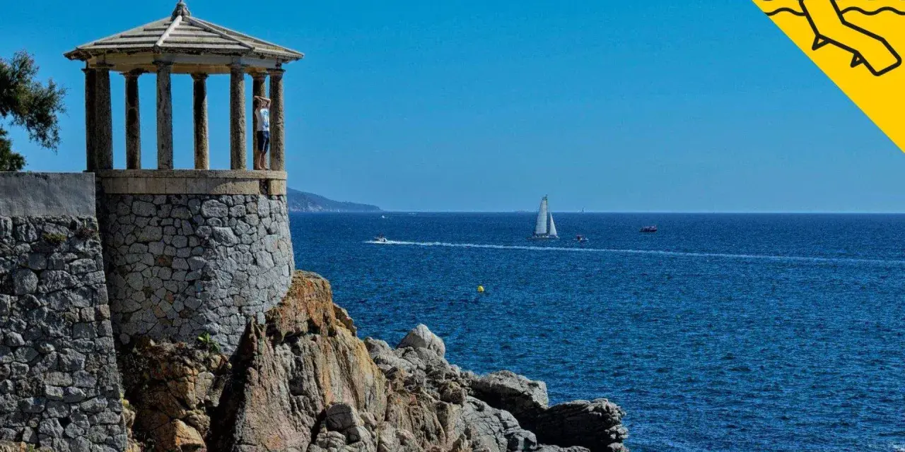 Un mirador de piedra con columnas se alza sobre rocas, con el mar azul y veleros al fondo. Un lugar perfecto para pasear por los **caminos de ronda**.