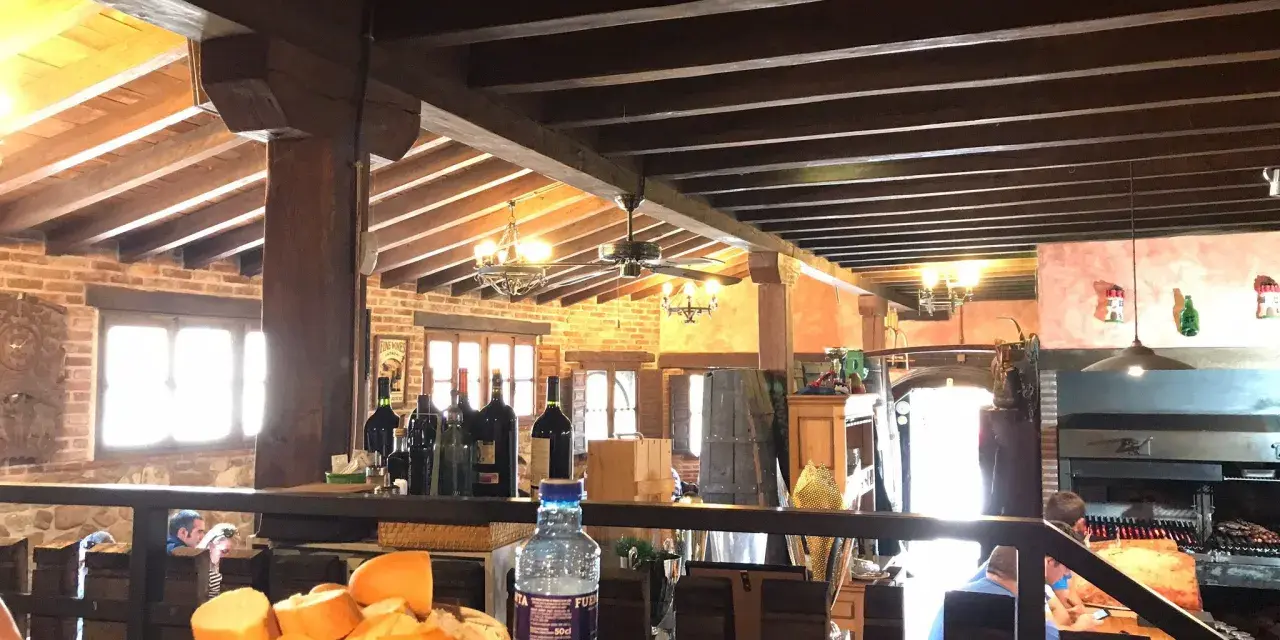 Interior rústico de restaurantes en Llanes, con vigas de madera, ladrillo y una parrilla a la vista. Cesta de pan y botella de agua en primer plano.