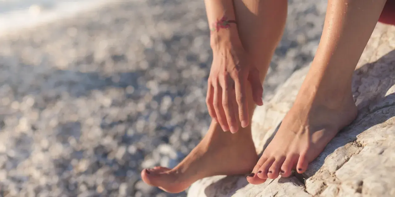 Pies descalzos sobre rocas en una playa nudista de Cadaqués. El sol brilla sobre la piel mojada y un pequeño tatuaje de flores adorna el brazo.