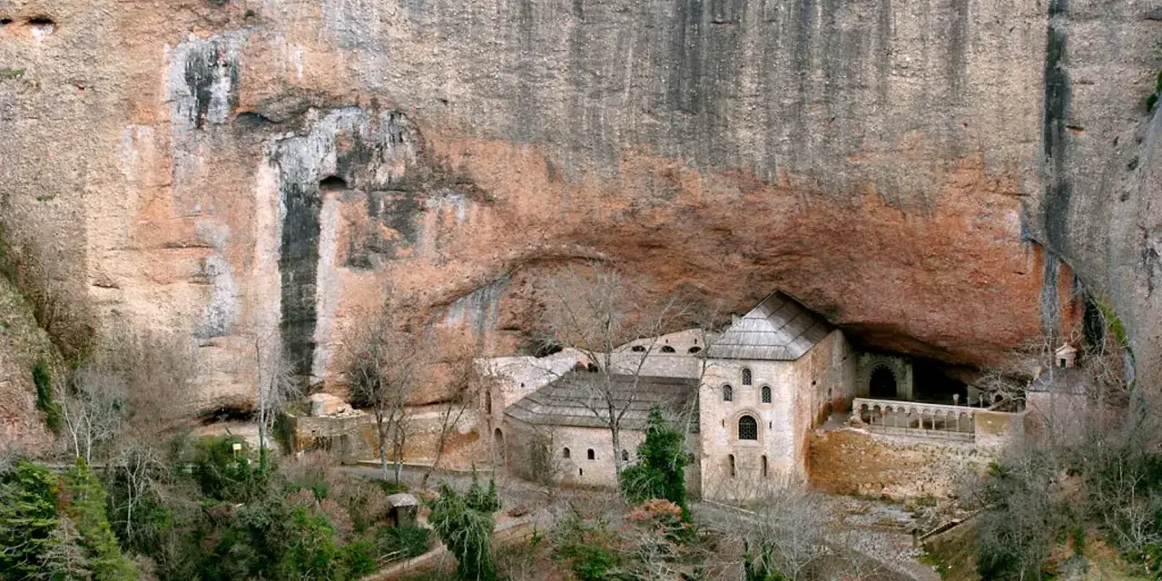 Monasterio de San Juan de la Peña, un impresionante conjunto arquitectónico excavado en la roca, con edificios de piedra y tejados a dos aguas bajo un gran saliente rocoso.