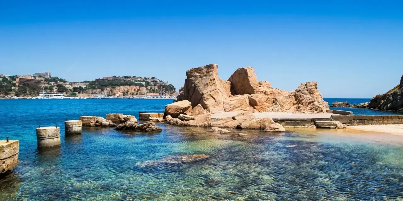 Un paseo marítimo en Sant Feliu de Guíxols, con rocas doradas y aguas cristalinas. El pueblo se alza en la colina.