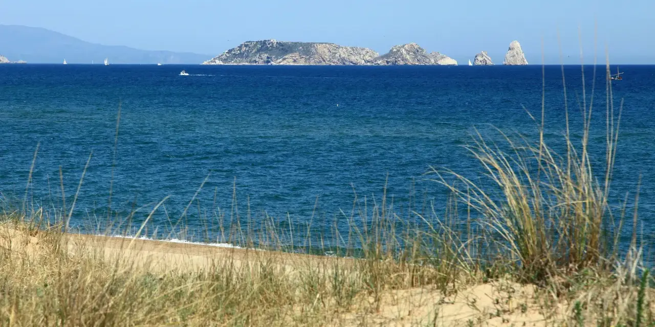 Dunas de hierba seca en primer plano, mar azul y un archipiélago rocoso en la distancia, evocando la belleza de la playa mas Pinell.