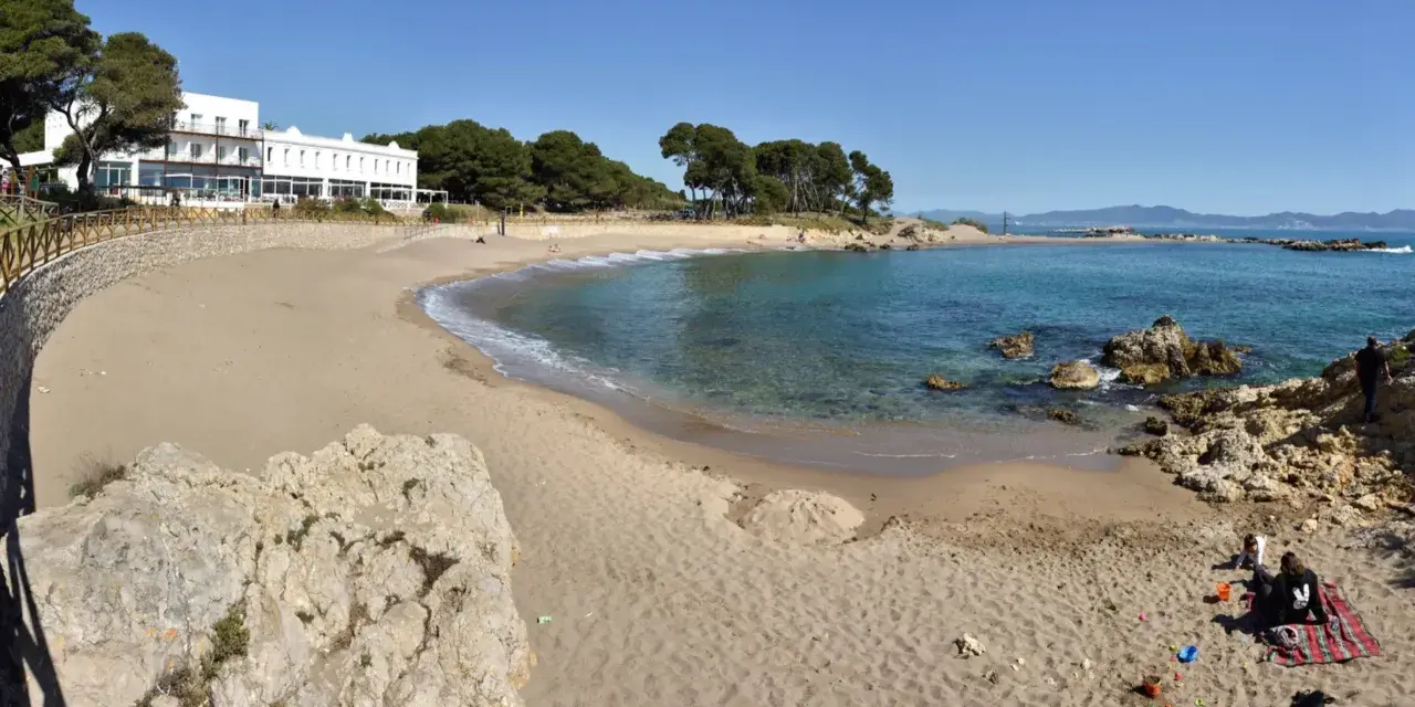 Platja Cala de la Creu: arena dorada, aguas turquesas y un hotel blanco con vistas al mar. Familias disfrutan del sol y juegan en la arena.