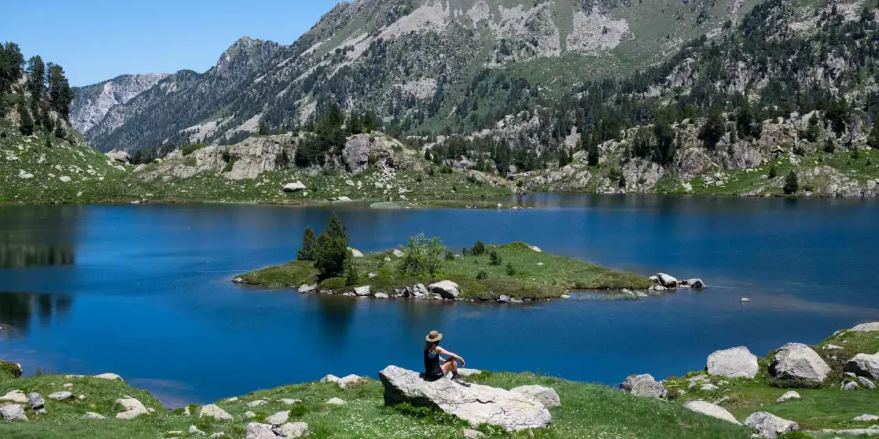 Persona sentada en roca contempla los lagos de Colomers en una ruta corta. Paisaje montañoso y azul intenso.