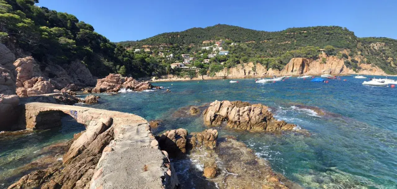 Un puente de piedra cruza aguas turquesas en la cala Canyet. Barcas flotan tranquilas mientras colinas verdes y casas blancas coronan el paisaje.