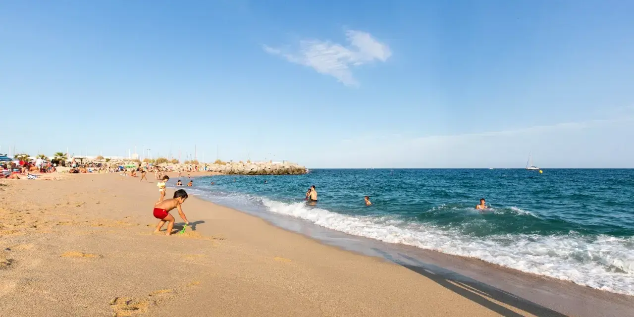 Un niño juega en la arena dorada de la playa de les barques, mientras otros disfrutan del mar azul y un velero navega en el horizonte.