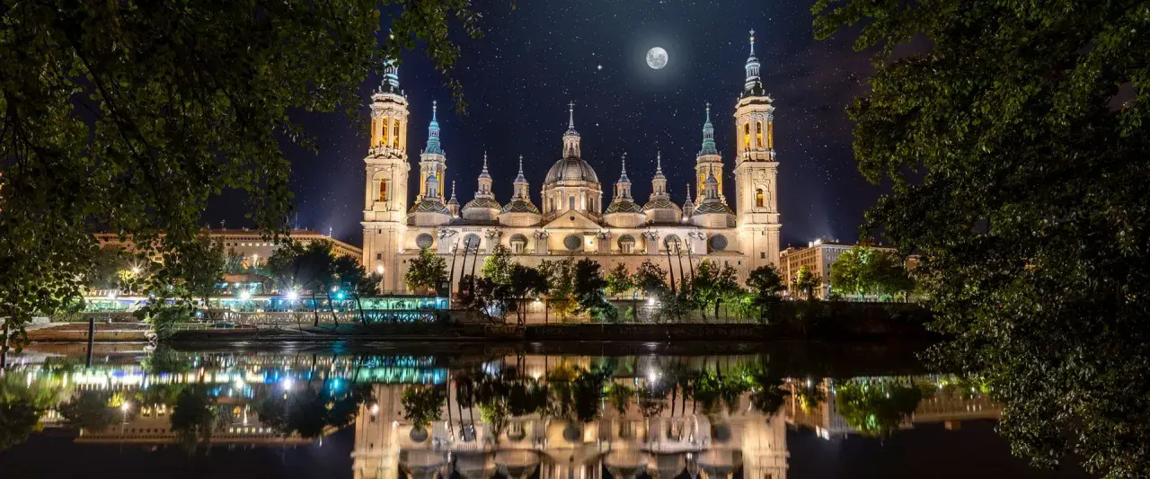 La majestuosa basílica del Pilar, iluminada bajo un cielo estrellado y la luna, se refleja en el río.