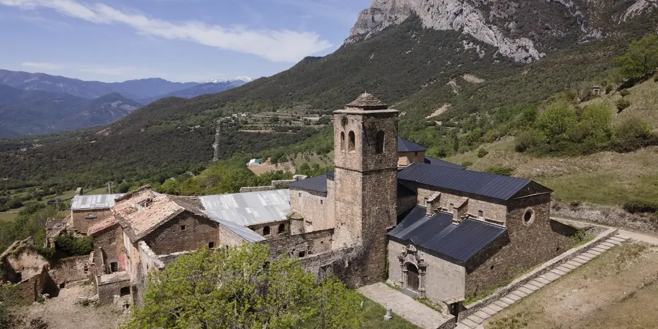 El monasterio de San Victorián, un conjunto de edificios de piedra con tejados oscuros, se alza majestuoso ante una imponente montaña rocosa.