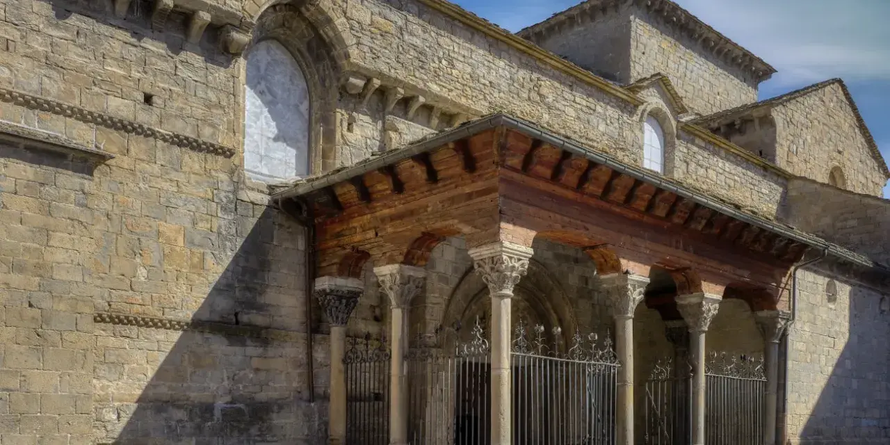 La **catedral de Jaca**, con su imponente arquitectura románica, se alza bajo un cielo azul. Un pórtico de madera y piedra protege la entrada.