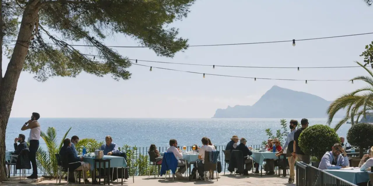 Gente disfrutando de una comida al aire libre con vistas al mar. Un lugar ideal donde comer en Altea, con mesas azules y un ambiente relajado bajo un árbol.