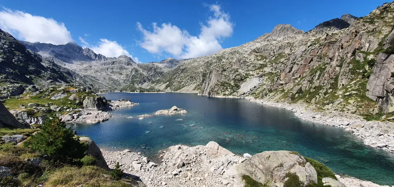 Lago de aguas cristalinas rodeado de montañas rocosas en el Vall de Boí. El cielo azul con nubes blancas contrasta con el verde de la vegetación.