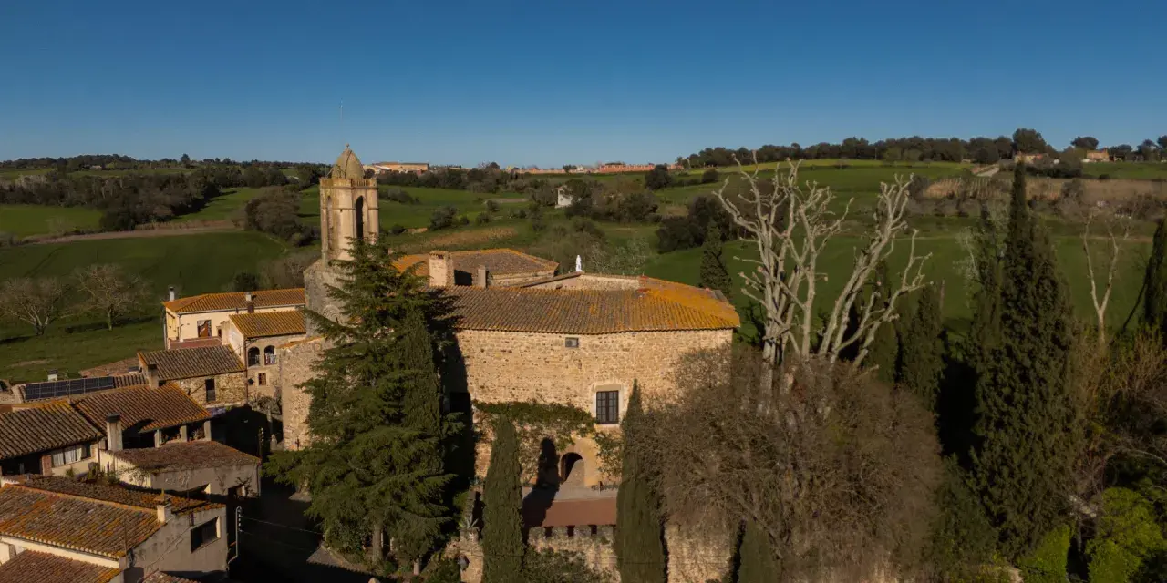 El castell de Gala, con su torre campanario, se alza entre tejados y vegetación, rodeado de verdes colinas bajo un cielo azul.