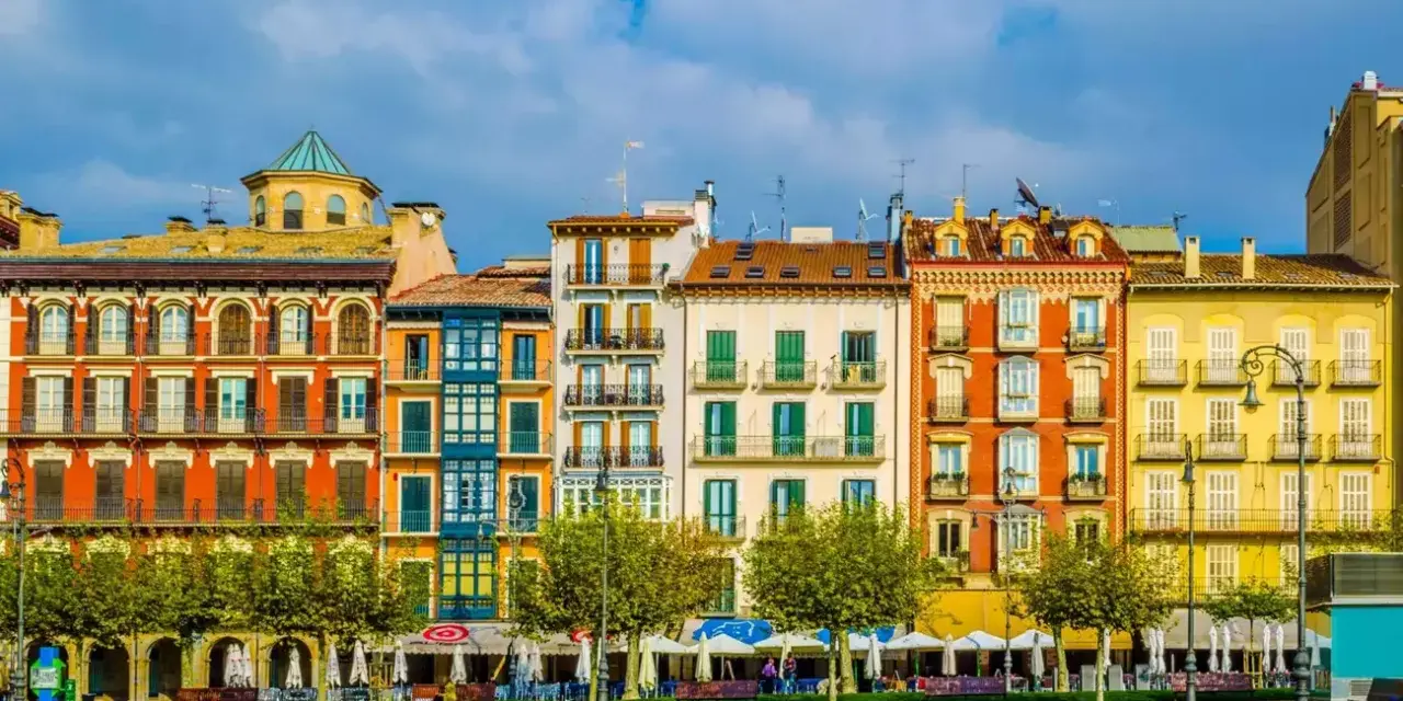 Fachada de edificios coloridos en una plaza, ideal para saber **qué ver en Pamplona**. Terrazas y árboles verdes bajo un cielo azul.