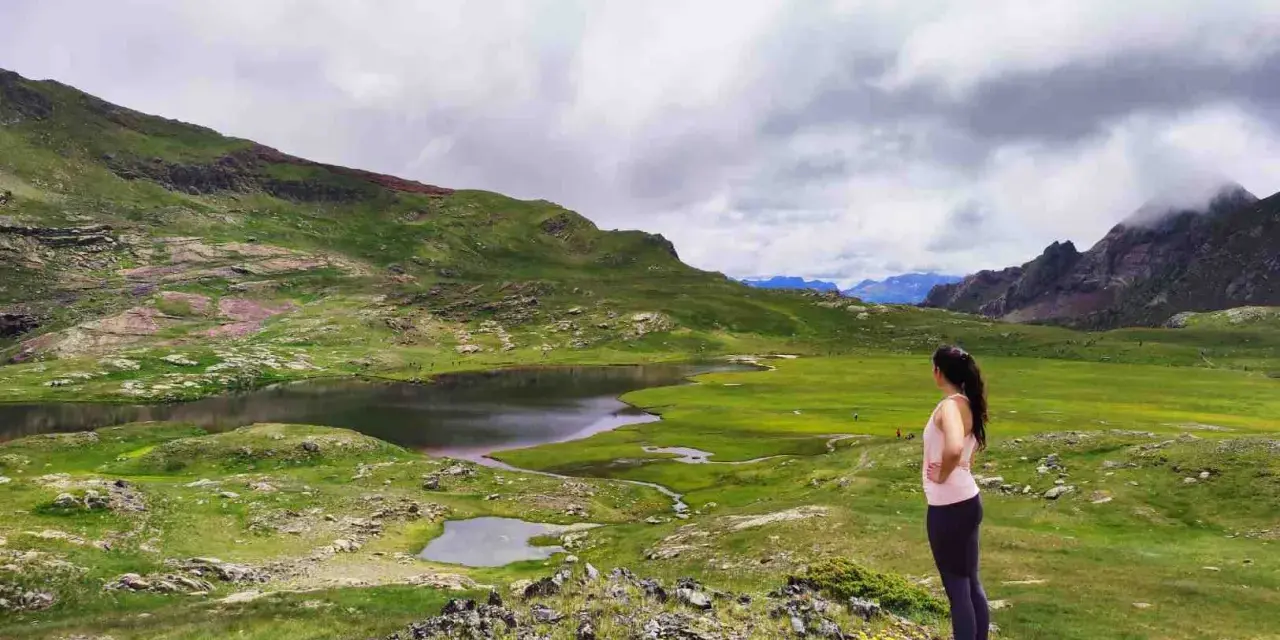 Mujer contempla los ibones del Valle de Tena, un paisaje de lagos y prados verdes bajo un cielo nublado.