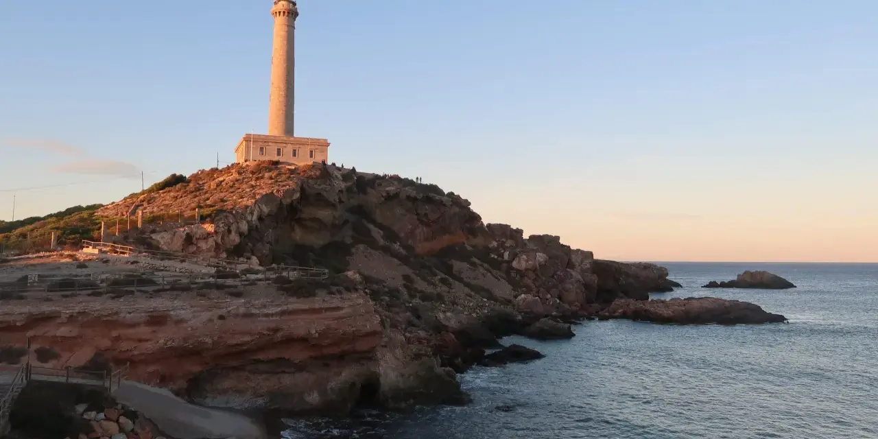 Faro sobre acantilados rojizos, con el mar azul y un cielo despejado. Un camino desciende hacia una pequeña cala roja.