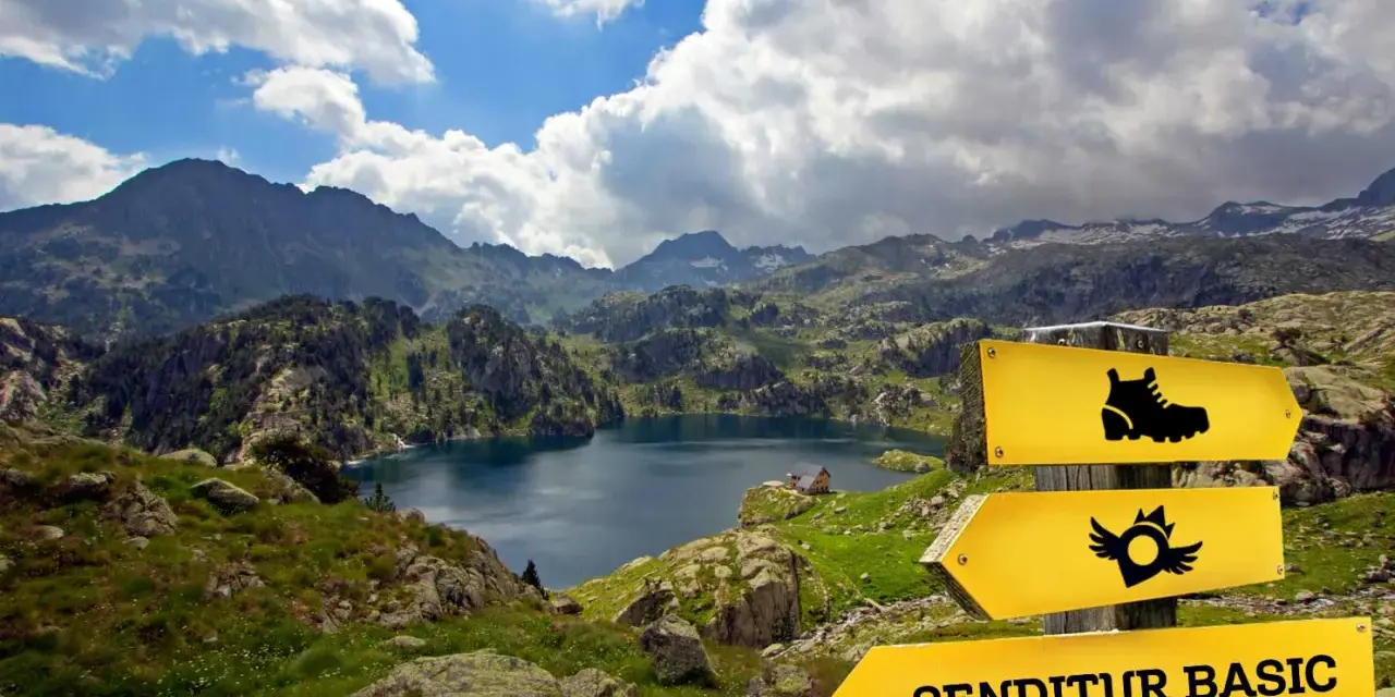 Paisaje de montaña con un lago azul, señalización de sendero y el letrero "SENDITUR BASIC", parte de la ruta de los 7 lagos Pirineos.