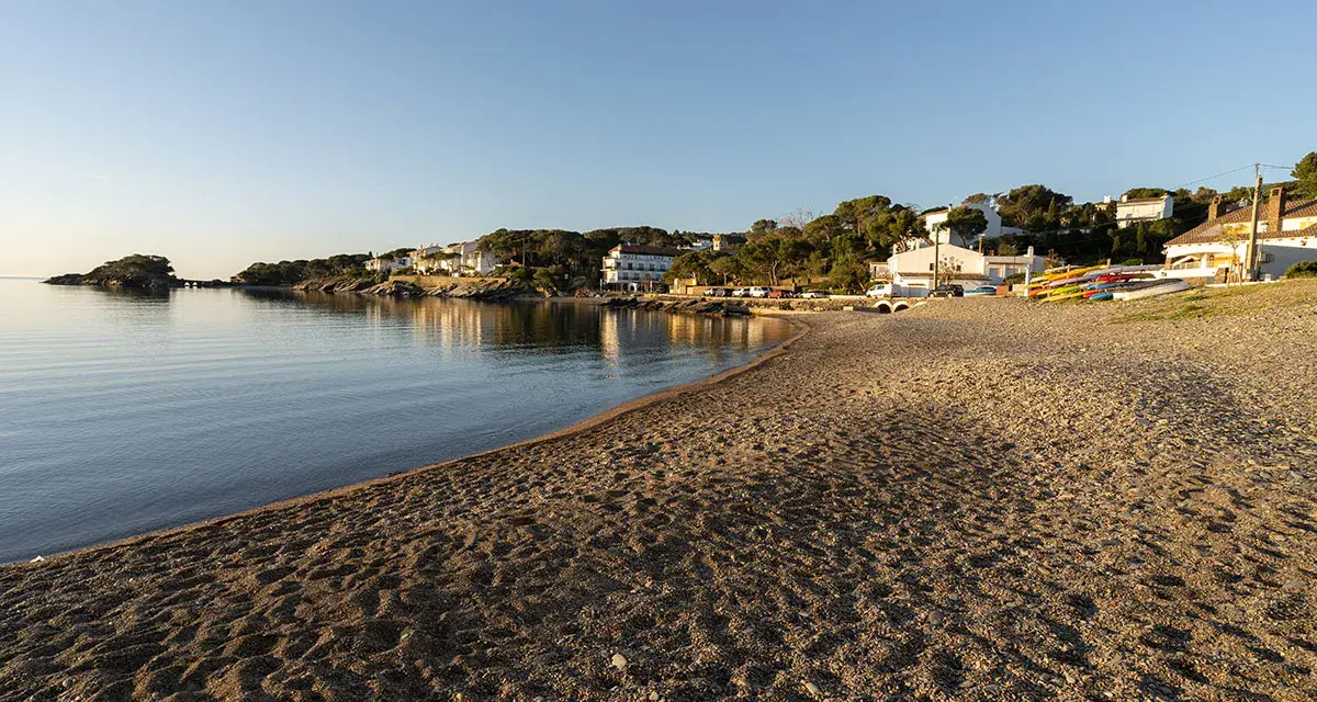 La platja gran de Cadaqués, con casas blancas y kayaks coloridos, se baña en la calma del mar al atardecer.