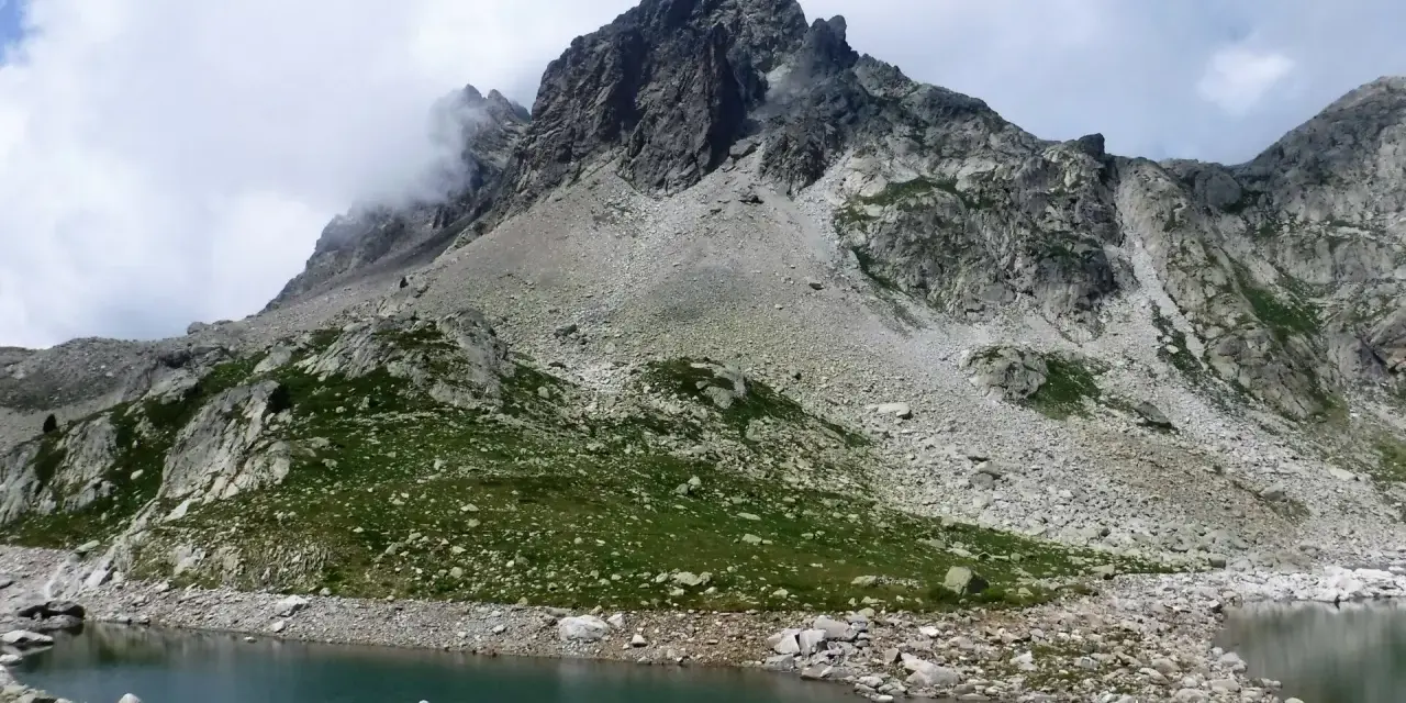 Paisaje montañoso con un lago de aguas tranquilas en primer plano. Las laderas están cubiertas de rocas y vegetación verde, con picos escarpados y nubes en el cielo. Los **ibones de Arriel** reflejan la majestuosidad del entorno.