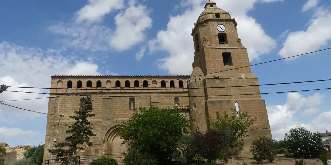 Iglesia de piedra con torre y reloj, rodeada de vegetación. Un día soleado en Alberuela de Tubo.