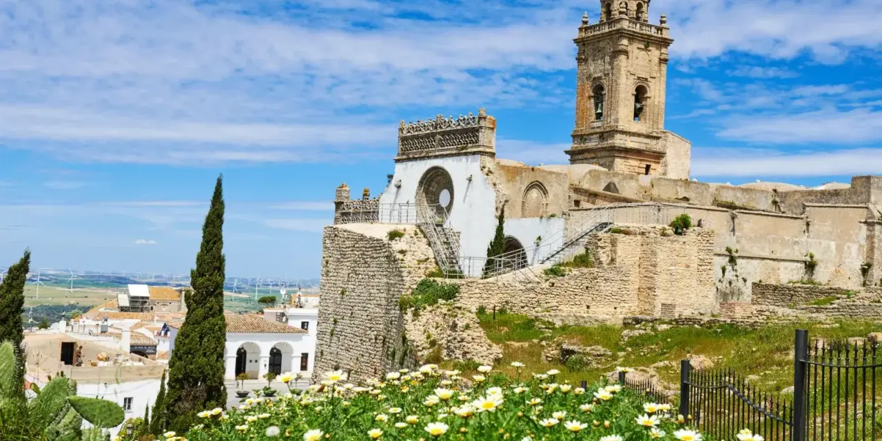 Descubre qué ver en Medina Sidonia: ruinas de un castillo, una iglesia con torre campanario y un campo de margaritas bajo un cielo azul.