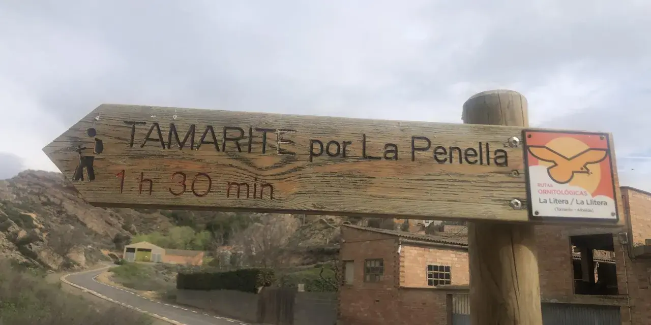 Señal de madera en albelda huesca: "Tamarite por La Penella, 1h 30 min". Al lado, señal de rutas ornitológicas.
