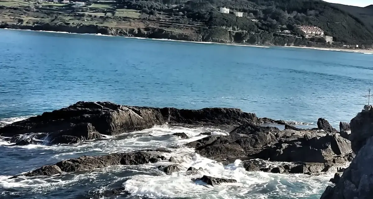 Olas rompiendo en rocas oscuras, con la costa verde y edificios de Mundaka al fondo. Un lugar perfecto para comer y disfrutar del paisaje.