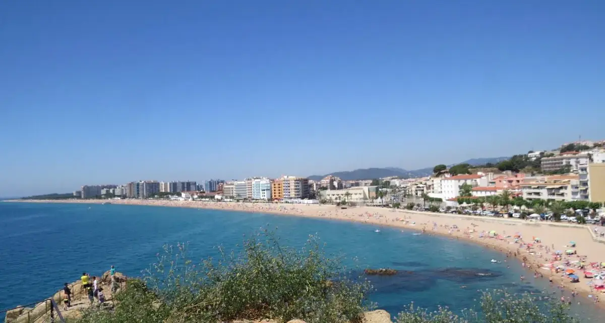 Vista panorámica de la playa S'Abanell en Blanes, con el mar azul intenso, arena dorada repleta de gente y edificios costeros bajo un cielo despejado.
