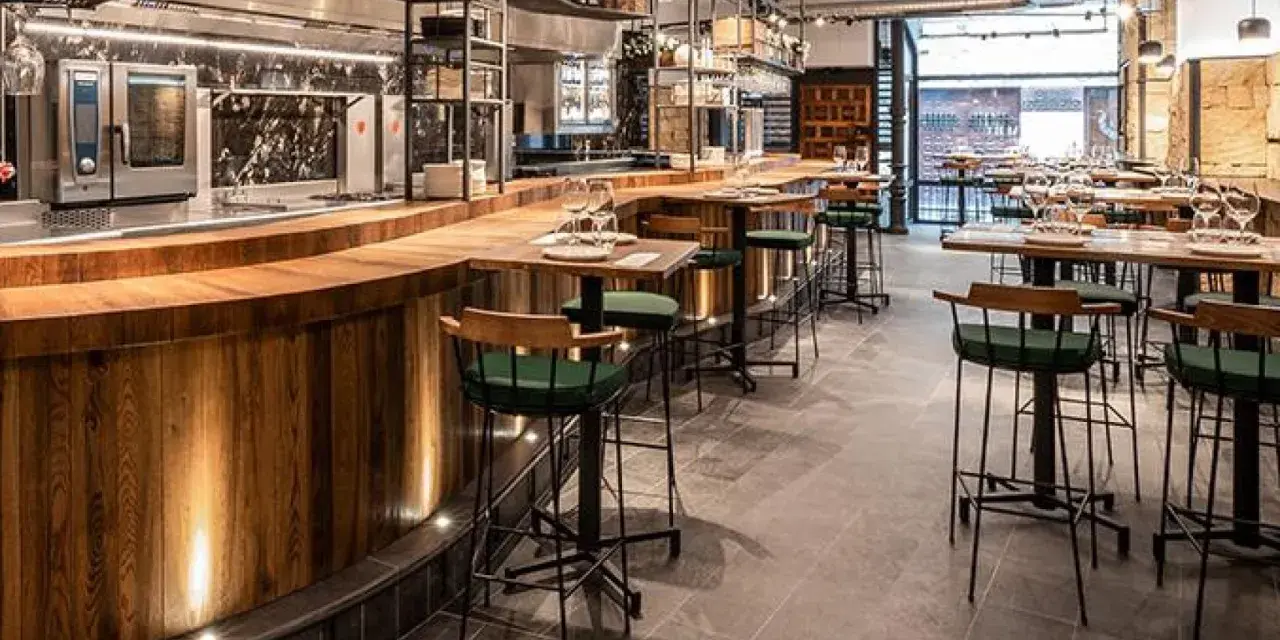 Bar moderno con taburetes verdes y mesas de madera. Un lugar ideal donde comer en Salamanca, con cocina a la vista y ambiente acogedor.