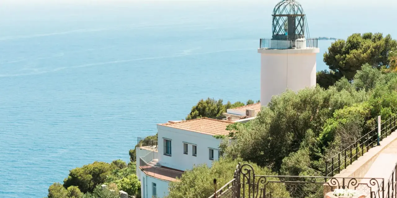 El faro de Sant Sebastià se alza sobre el mar azul, rodeado de vegetación exuberante.