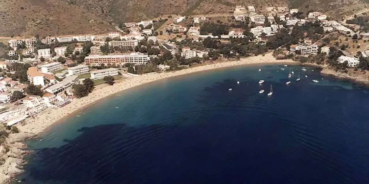 Vista aérea de la playa Almadrava en Roses, con edificios blancos y barcos anclados en aguas azules.