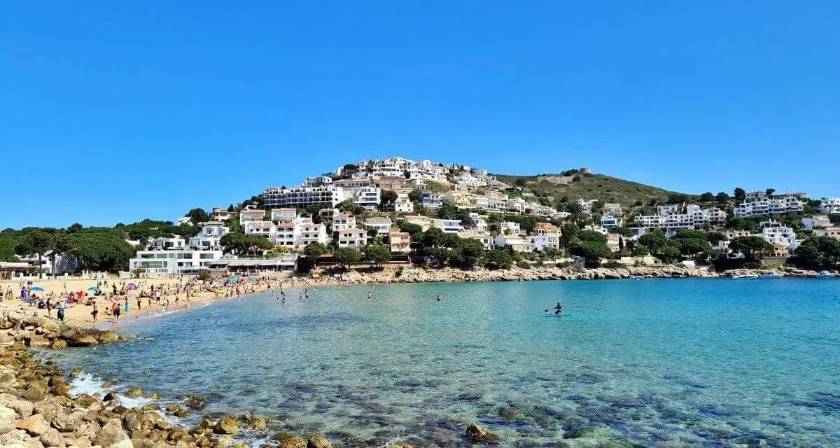 Vista de la platja de Montgó, con gente disfrutando del sol y el mar turquesa, y casas blancas escalonadas en la colina.
