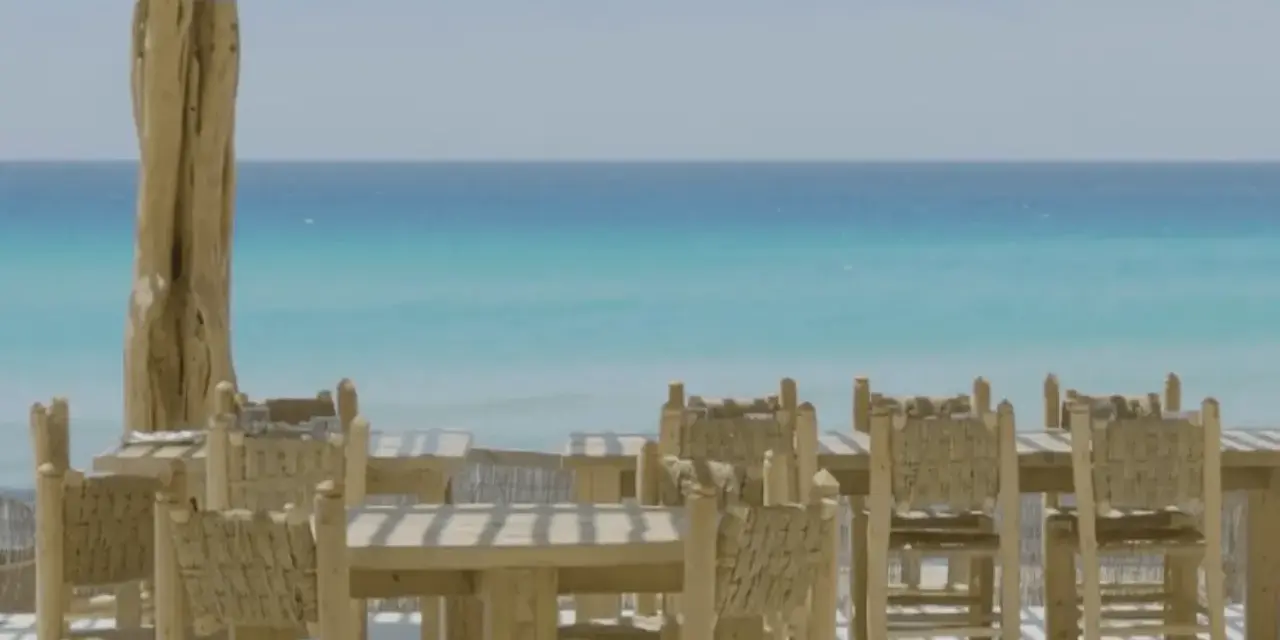 Mesas y sillas de madera rústica en la terraza de restaurantes en Formentera, con el mar turquesa de fondo.