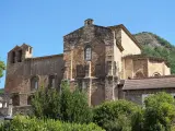El **monasterio de Siresa**, con su imponente arquitectura de piedra y campanario, se alza bajo un cielo azul, rodeado de vegetación exuberante.
