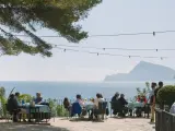 Gente disfrutando de una comida al aire libre con vistas al mar. Un lugar ideal donde comer en Altea, con mesas azules y un ambiente relajado bajo un árbol.