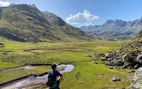 Un excursionista contempla un vasto valle de hecho, con un río serpenteante y montañas majestuosas bajo un cielo azul.