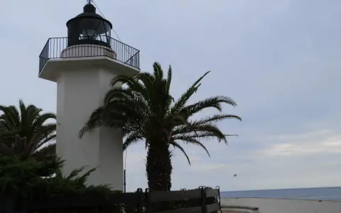 El faro de Palamós, blanco y esbelto, se alza junto a palmeras y el mar azul.