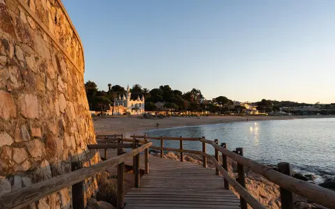 Pasarela de madera hacia la platja Sant Pol, Sant Feliu de Guíxols. Edificios blancos y palmeras bordean la arena dorada bajo un cielo azul.