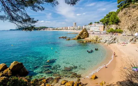 Una caleta en la Costa Brava con aguas cristalinas, buceadores explorando el fondo rocoso y una playa con gente disfrutando del sol.
