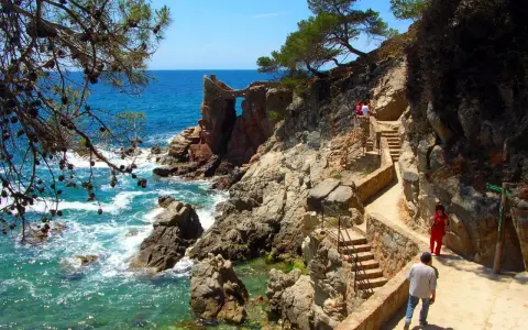 Un camino de ronda con escaleras de piedra serpentea por la costa rocosa. El mar turquesa rompe contra las rocas.