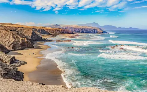 Paisaje de las playas de Fuerteventura con acantilados rocosos, arena dorada y el mar turquesa con olas rompiendo.