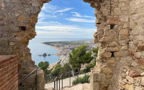 Vista desde el **castillo de Blanes** hacia la bahía y la ciudad. El mar azul contrasta con las ruinas de piedra y el cielo nublado.