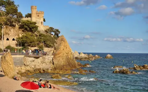 Castillo medieval sobre acantilados rocosos en sa caleta Lloret de Mar. Playa con gente y mar azul.