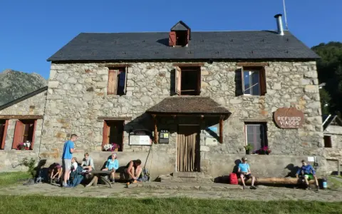 Grupo de excursionistas descansa frente al Refugio Viados, parte de la ruta de los tres refugios. Montañas y cielo azul al fondo.