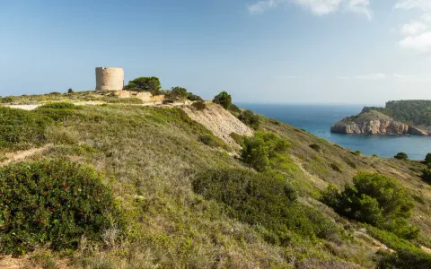 La torre de Montgó se alza sobre una colina verde, vigilando el mar azul y una bahía rocosa.