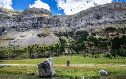 Senderista camina por un prado verde con imponentes montañas de **3000 del Pirineo** al fondo.