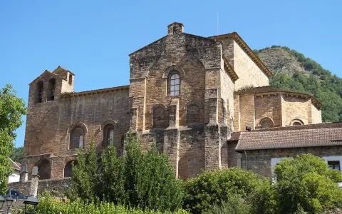 El **monasterio de Siresa**, con su imponente arquitectura de piedra y campanario, se alza bajo un cielo azul, rodeado de vegetación exuberante.