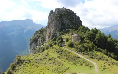 Pequeña ermita de piedra con tejado cónico, rodeada de vegetación y un sendero que sube por la ladera. Las ermitas de Tella se alzan majestuosas.