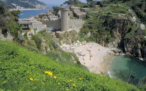 Vista de las calas de Tossa de Mar, con un castillo medieval y una playa con gente disfrutando del sol y el mar.