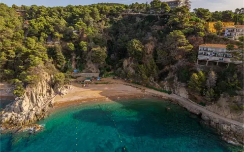 Bahía de Cala Bona, Barcelona, con aguas turquesas, arena dorada y acantilados verdes. Casas en la ladera.